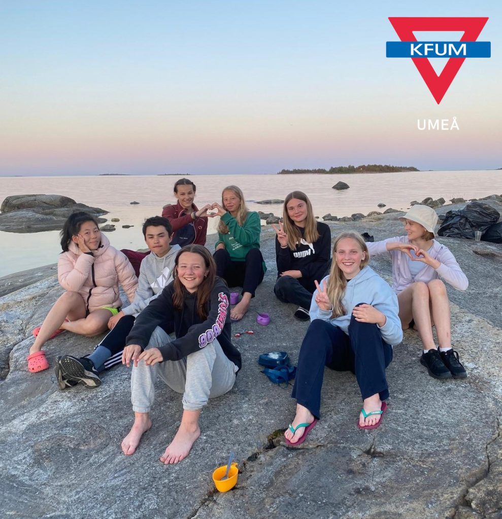 Bilden visar en grupp deltagare och ledare på klipporna vid Norrbyskärs lägergård. Deltagarna ser glada ut och gör hjärtan med händerna.